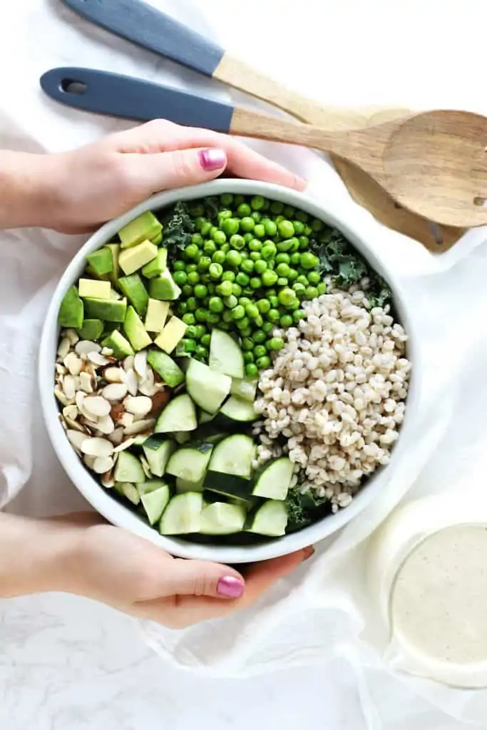 Two hands holding green grain summer salad in white bowl.