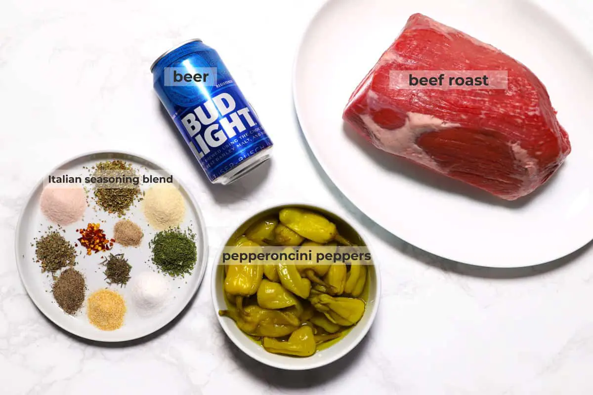 Beef roast, pepperoncini peppers, can of beer and spices to make Italian seasoning blend on a marble surface. 