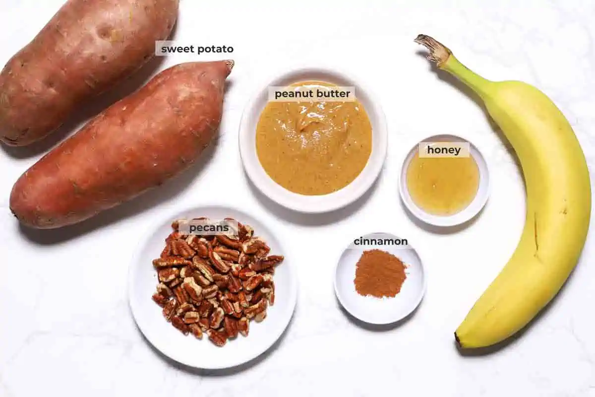 Sweet potatoes, peanut butter, pecans, cinnamon, honey and a banana on a marble counter. 