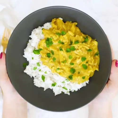 Two hands holding a dark grey bowl of honey curry chicken and coconut rice.