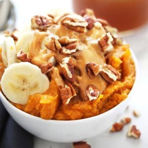 Sweet potatoes with banana slices, peanut butter and walnuts in a white bowl on grey napkin.