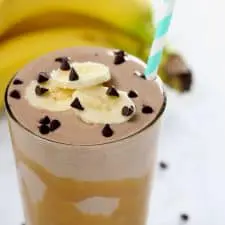 Chocolate smoothie with banana slices in clear glass on white surface.