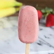Pink popsicles on marble surface next to strawberries and bananas.