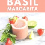 Pink margarita next to strawberries, basil and limes.