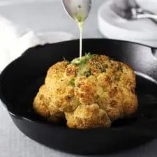 Whole head of roasted cauliflower in cast iron pan with butter being poured on top.